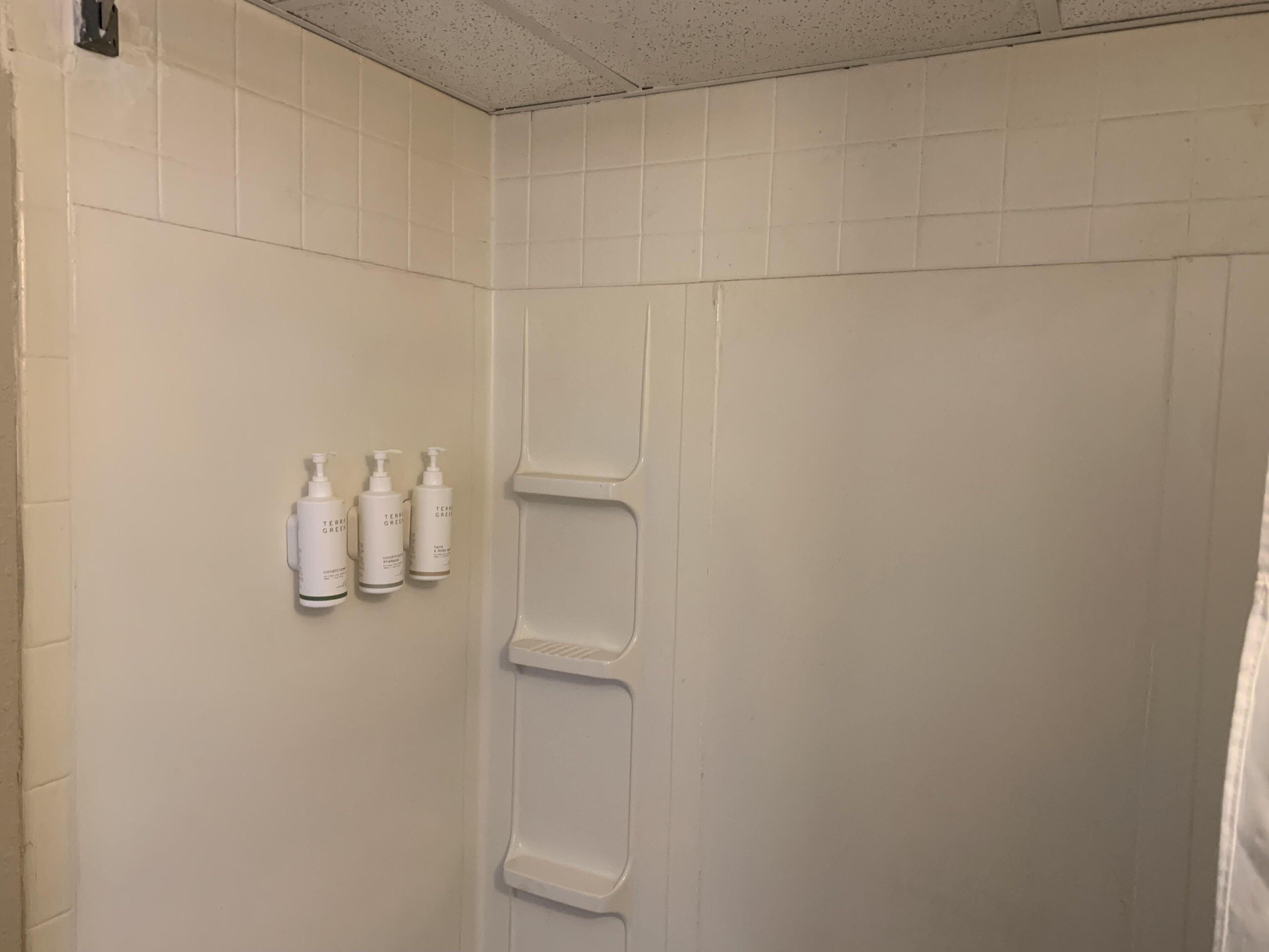 Baño con paredes de azulejos blancos y zona de ducha. Hay tres botellas de champú 'Terri Green' en la pared.