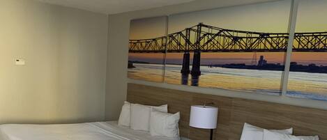 Quarto de hotel com duas camas, uma mesa de cabeceira com abajur e uma grande pintura de uma ponte ao pôr do sol.