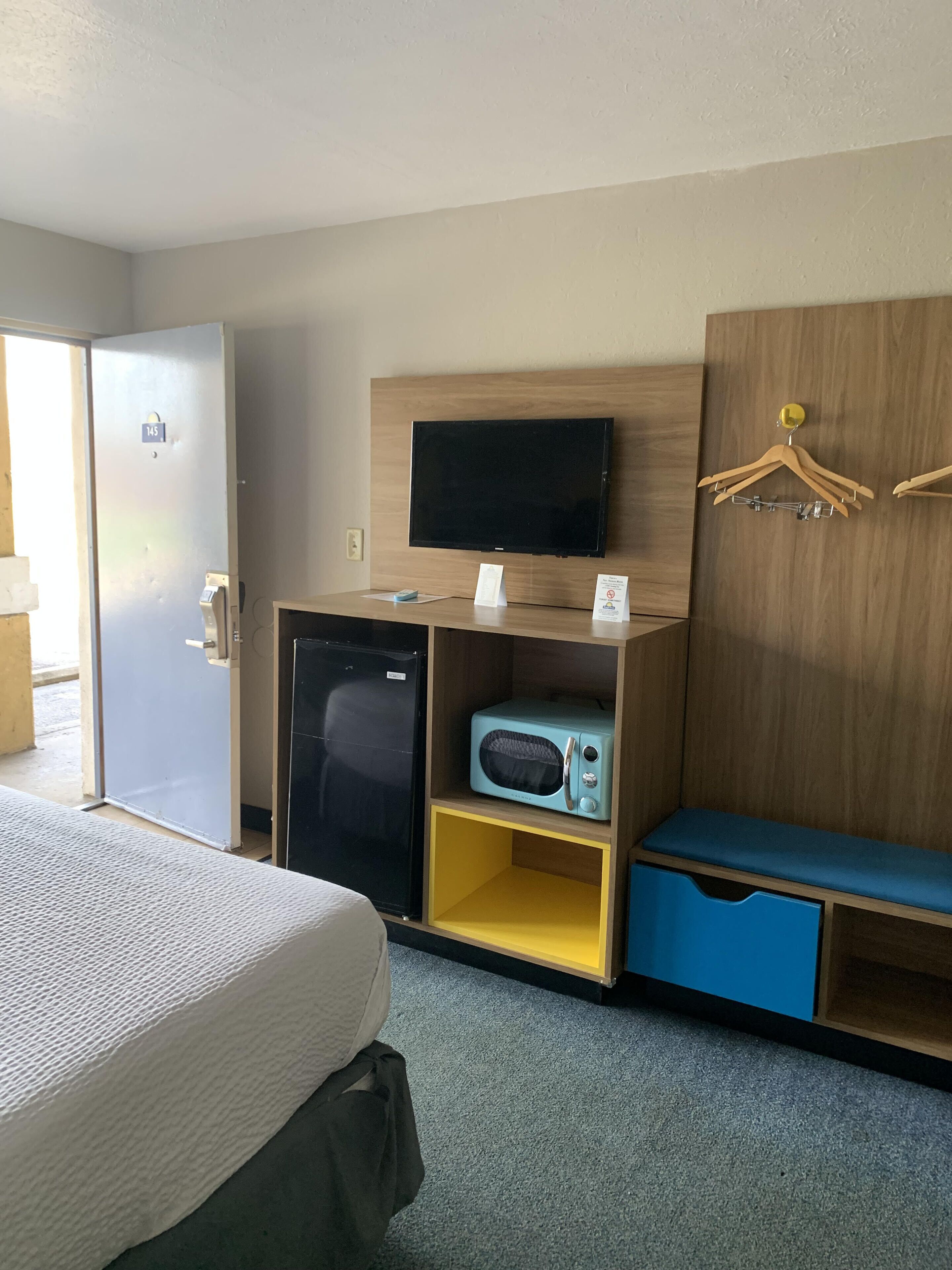 Quarto de hotel com uma cama, um pequeno frigorífico, um micro-ondas, uma televisão e um guarda-roupa de madeira com um banco azul.