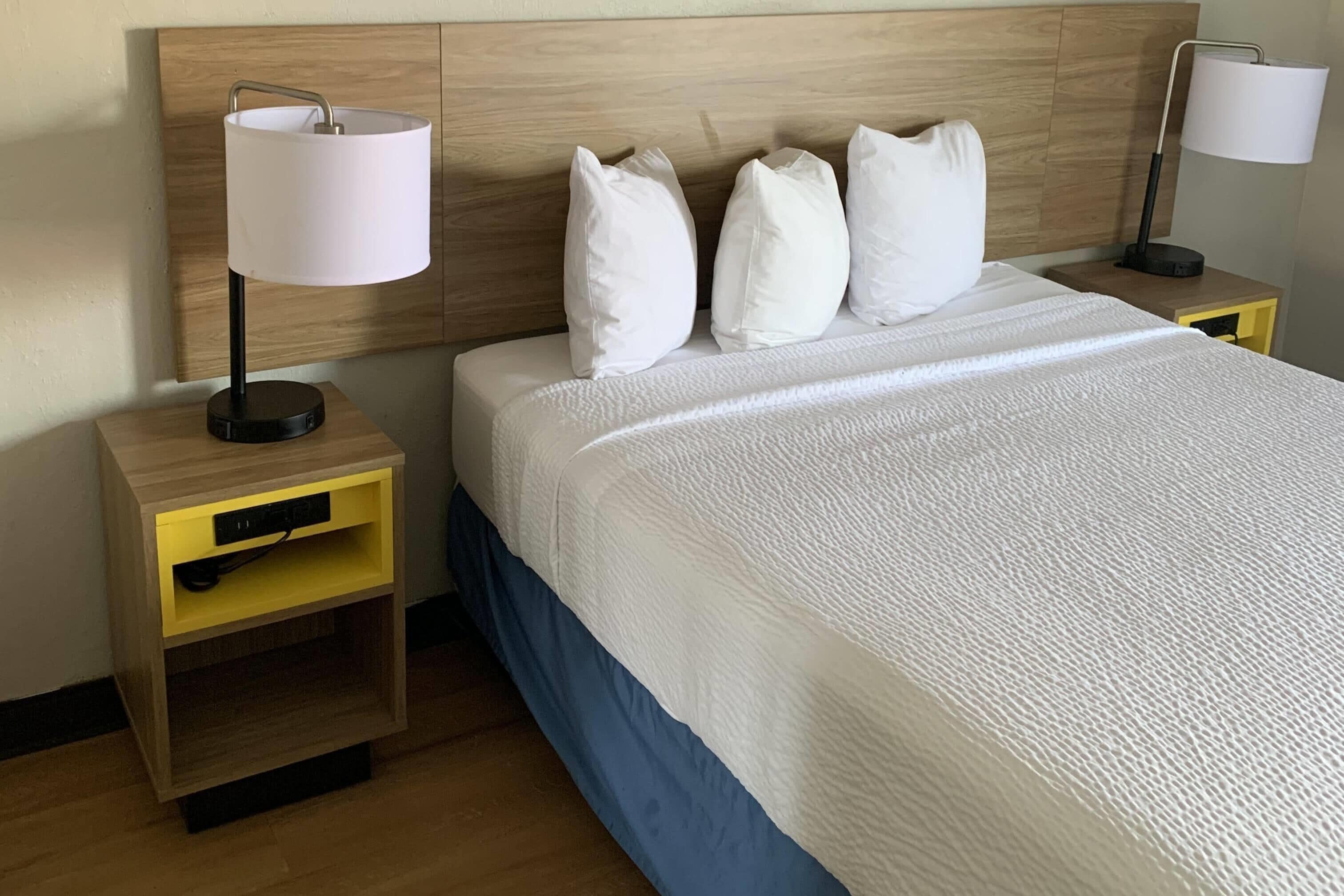 Habitación de hotel con cabecera de madera, cama con ropa de cama blanca, dos mesitas de noche con lámparas y un tocador amarillo con cajón.