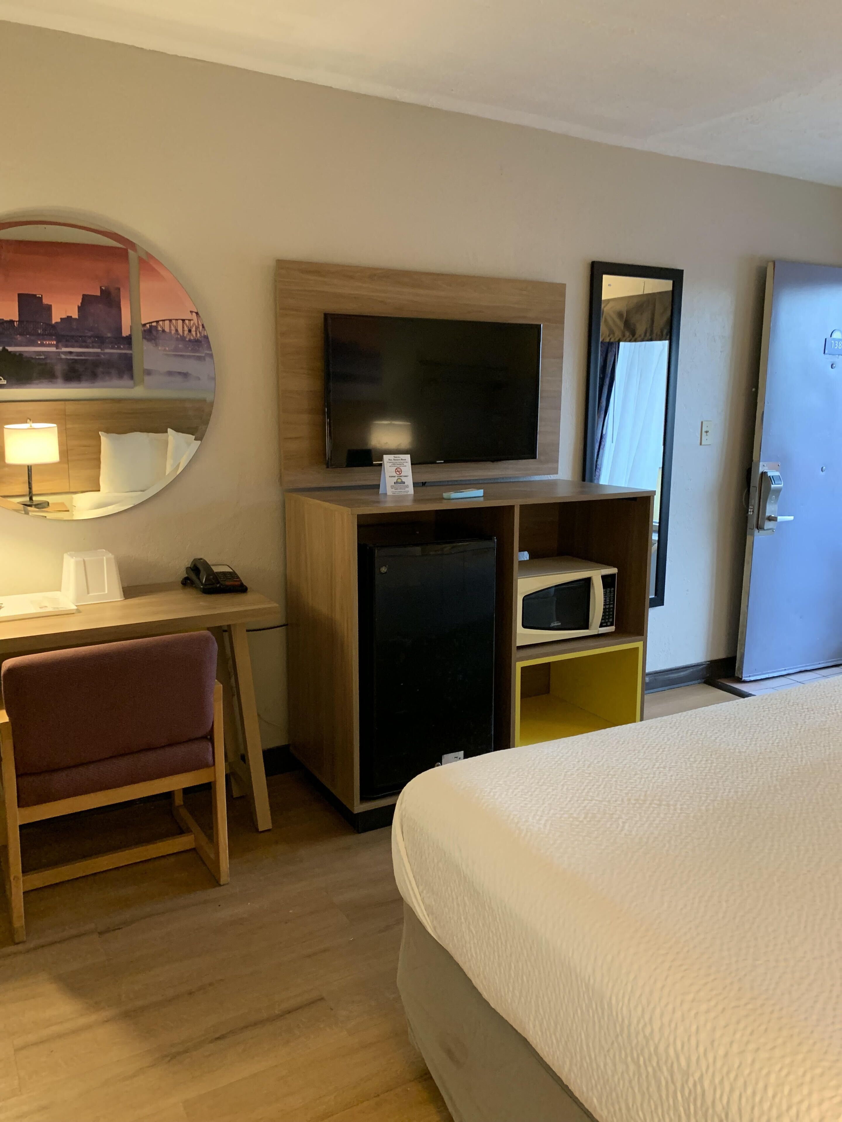 Ett hotellrum med en säng, ett skrivbord med en stol, en tv, ett kylskåp, en mikrovågsugn och en spegel.