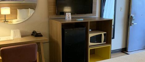 Ett hotellrum med en säng, ett skrivbord med en stol, en tv, ett kylskåp, en mikrovågsugn och en spegel.