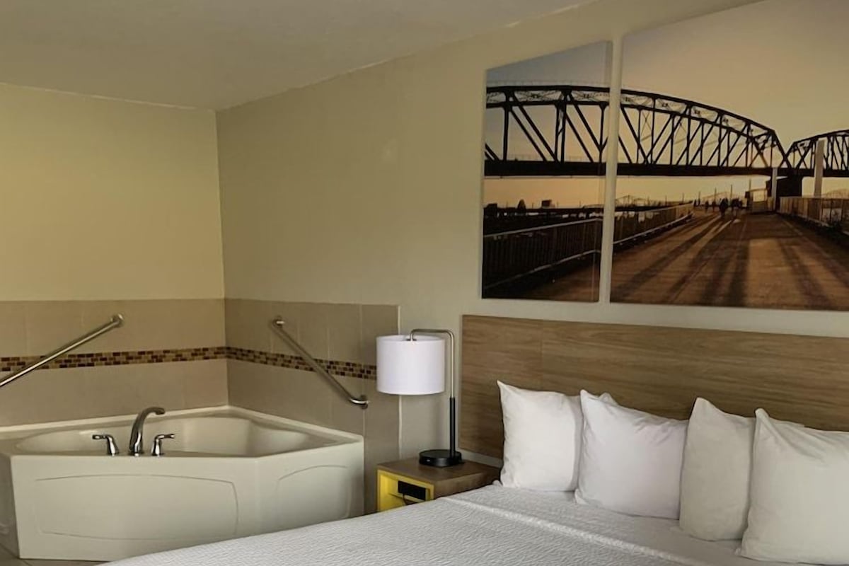 Habitación de hotel con una cama grande, una bañera independiente, una mesita de noche con lámpara y un cuadro colgado en la pared de un puente.
