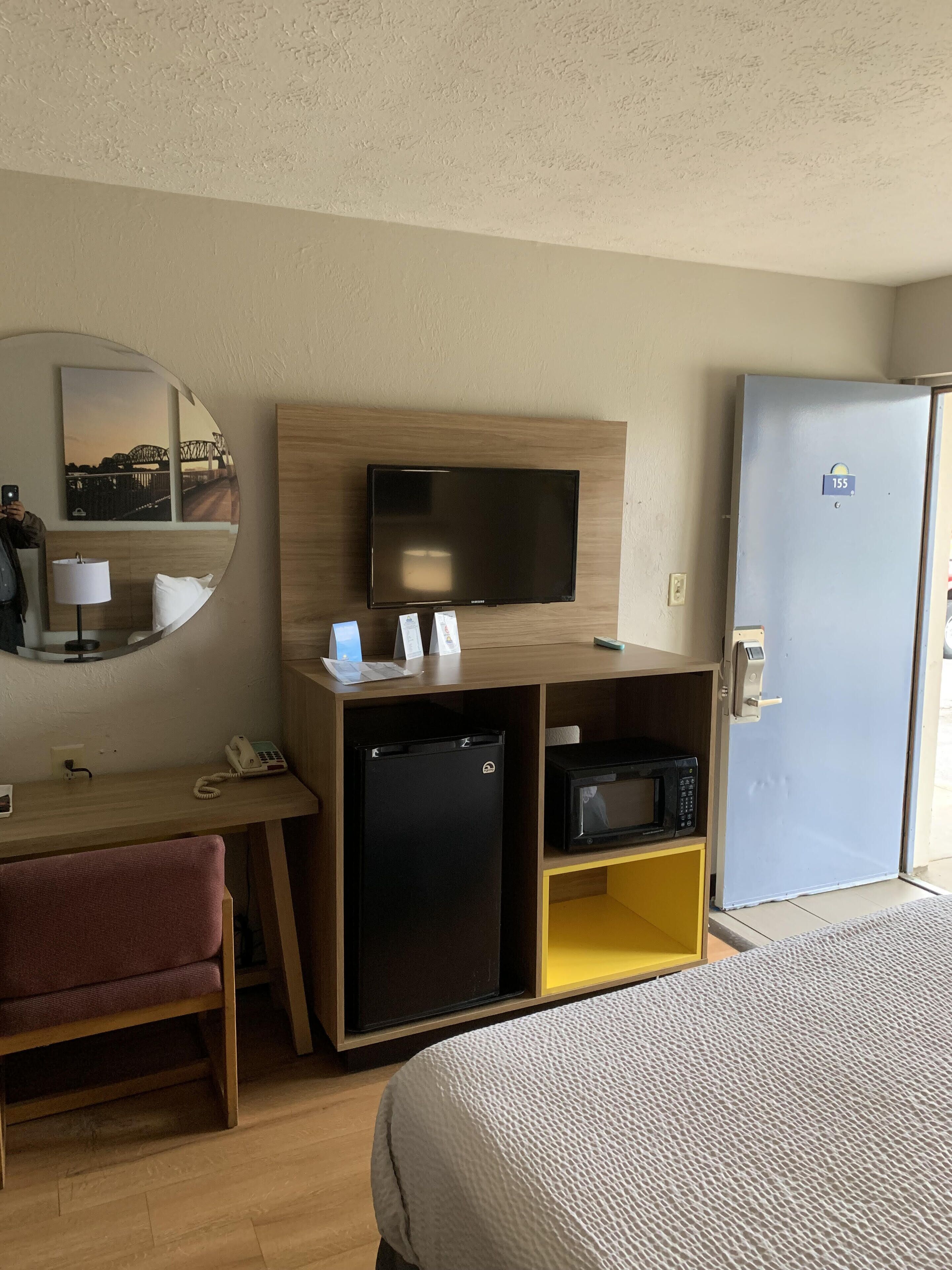 Una habitación de hotel con cama, escritorio con teléfono, refrigerador pequeño, microondas y televisor sobre un mueble de madera.