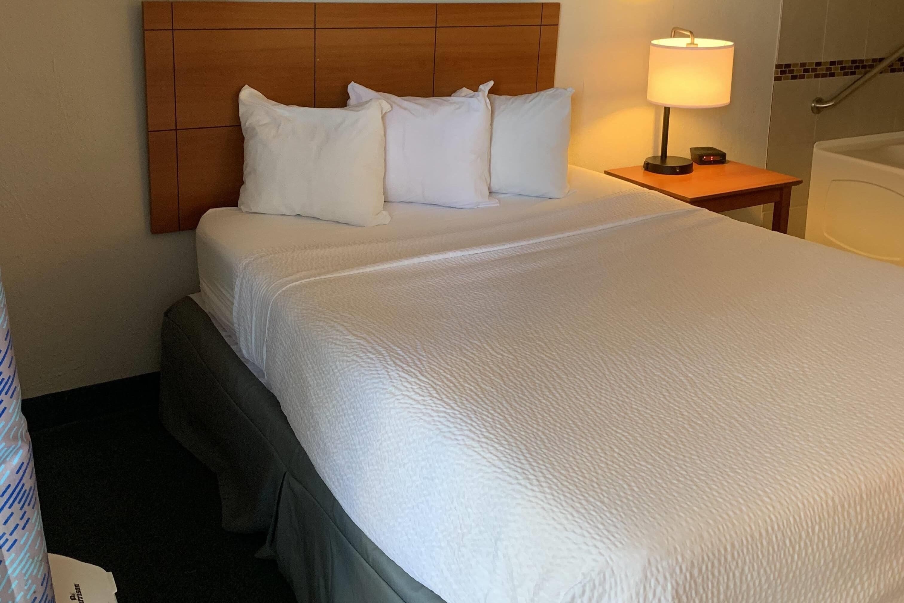 A hotel room with a large bed, a wooden headboard, a bedside table with a lamp, and a bathtub.