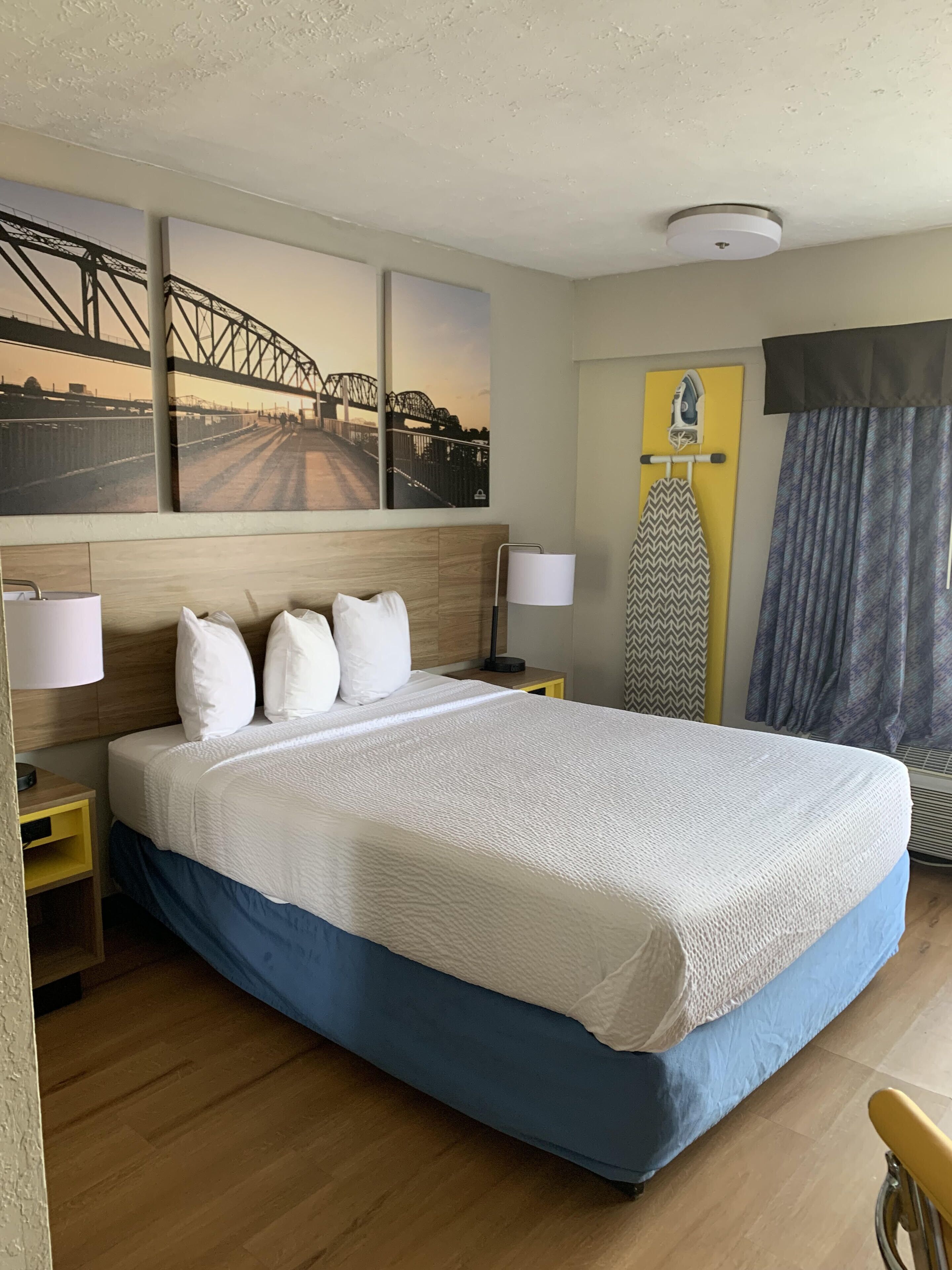 A hotel room with a large bed, wooden headboard, and a painting of a bridge. There is a nightstand with a lamp and an ironing board with an iron.