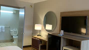 A hotel room with a bed, a desk with a lamp, a television, a microwave, and a bathroom visible through an open door.