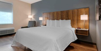 Habitación de hotel con una cama grande, cabecera de madera, dos mesitas de noche con lámparas, una silla y una ventana con persianas.