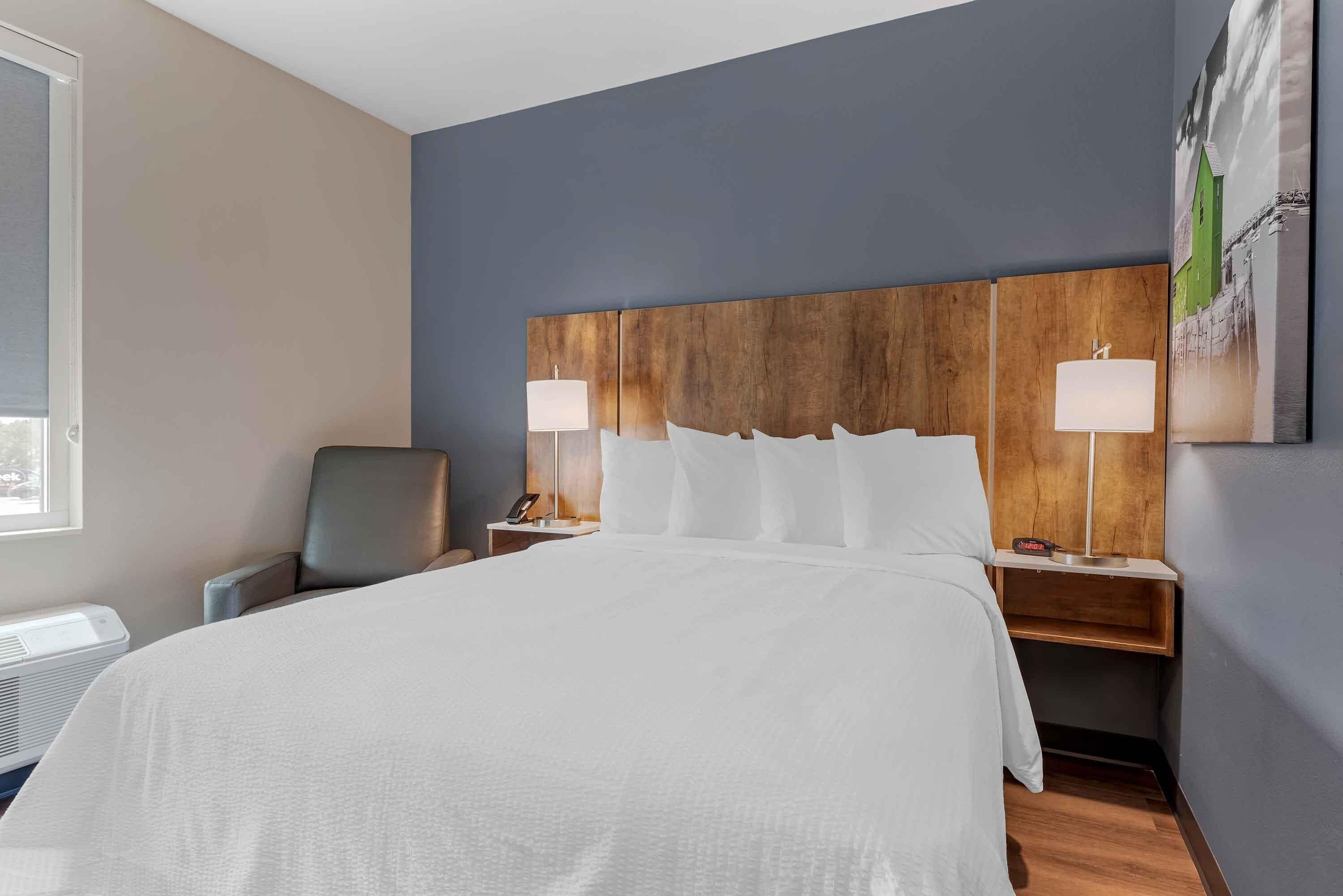 Una habitación de hotel con una cama grande, dos mesitas de noche con lámparas, una silla y una obra de arte colgada en la pared.