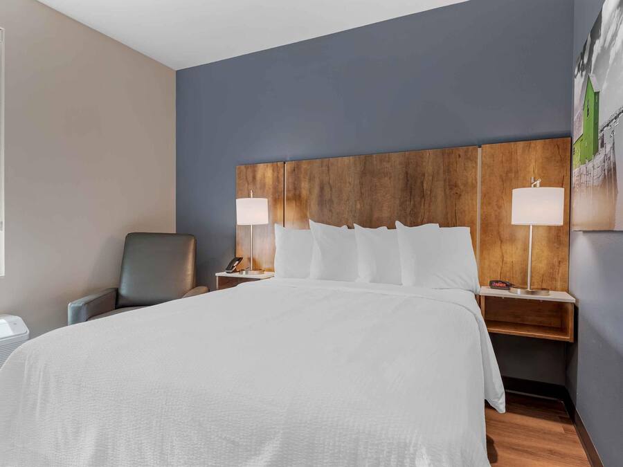 Una habitación de hotel con una cama grande, dos mesitas de noche con lámparas, una silla y una obra de arte colgada en la pared.