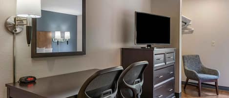 A hotel room with a desk, two chairs, a television, a mirror, and a wall-mounted lamp.