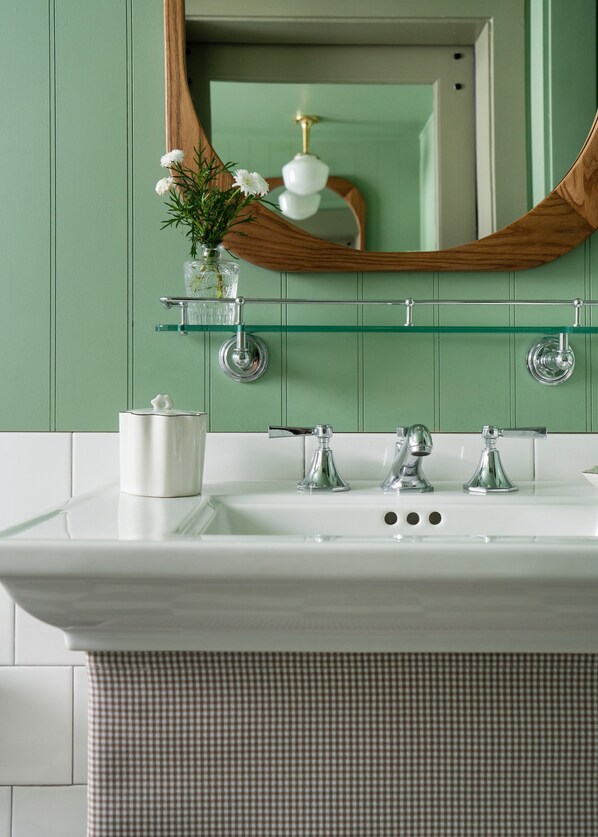 A bathroom with a white sink, three faucets, a glass shelf with a vase of flowers, and a wooden-framed mirror.