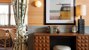 A room with a wooden console table, a framed picture of a beach scene, a lamp, and a decorative shelf with books and figurines.