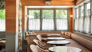 A cozy seating area with wicker chairs and tables, large windows with curtains, and a wooden ceiling.