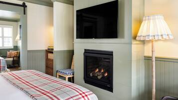 A cozy room with a fireplace, a television mounted above it, a plaid blanket on the bed, and a lamp beside the chair.