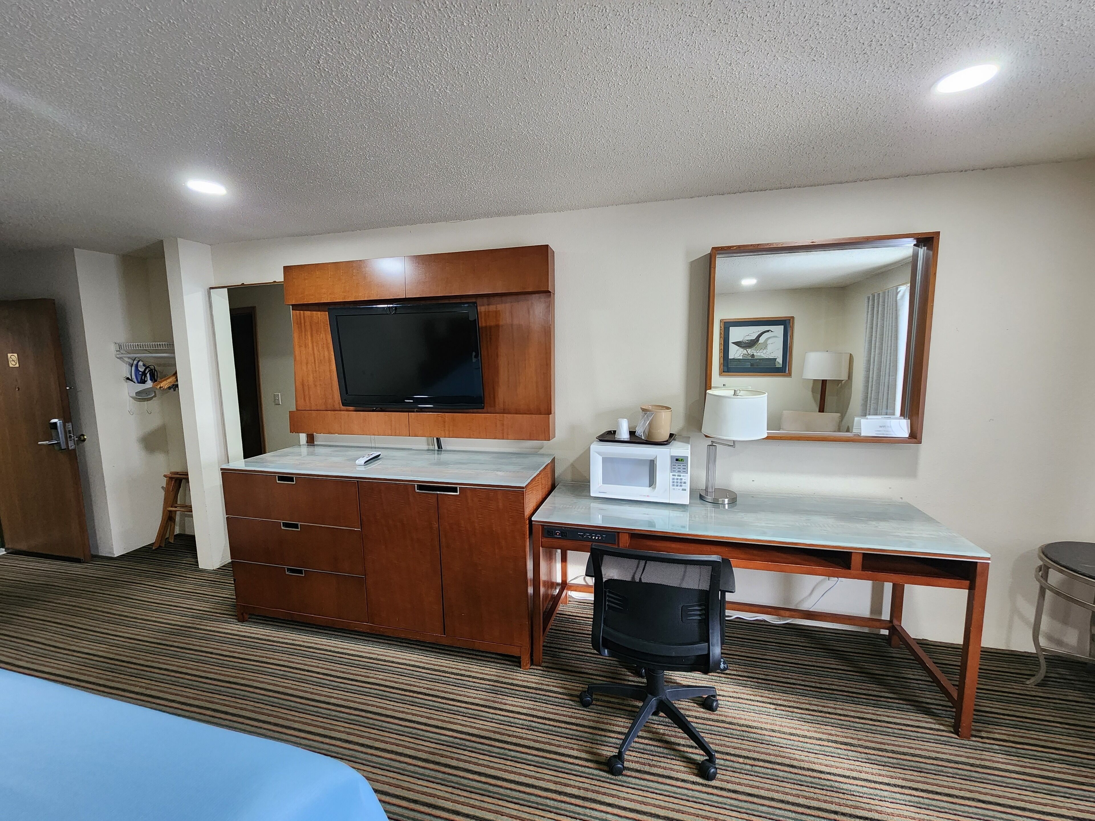 Une chambre d’hôtel avec un bureau en bois, un téléviseur fixé au-dessus, un four à micro-ondes, une chaise et un lit recouvert d’une couverture bleue.