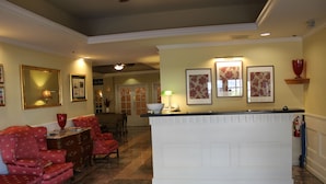 A hotel lobby with a reception desk, red upholstered chairs, a white counter, framed floral artwork, and a fire extinguisher. - Hotel Chester (Starkville)