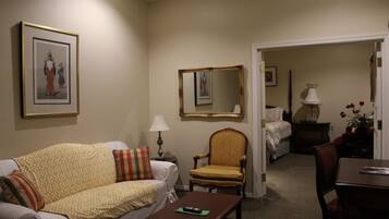 A hotel room with a sofa, a chair, a table, a lamp, a mirror, and a bed in the background.