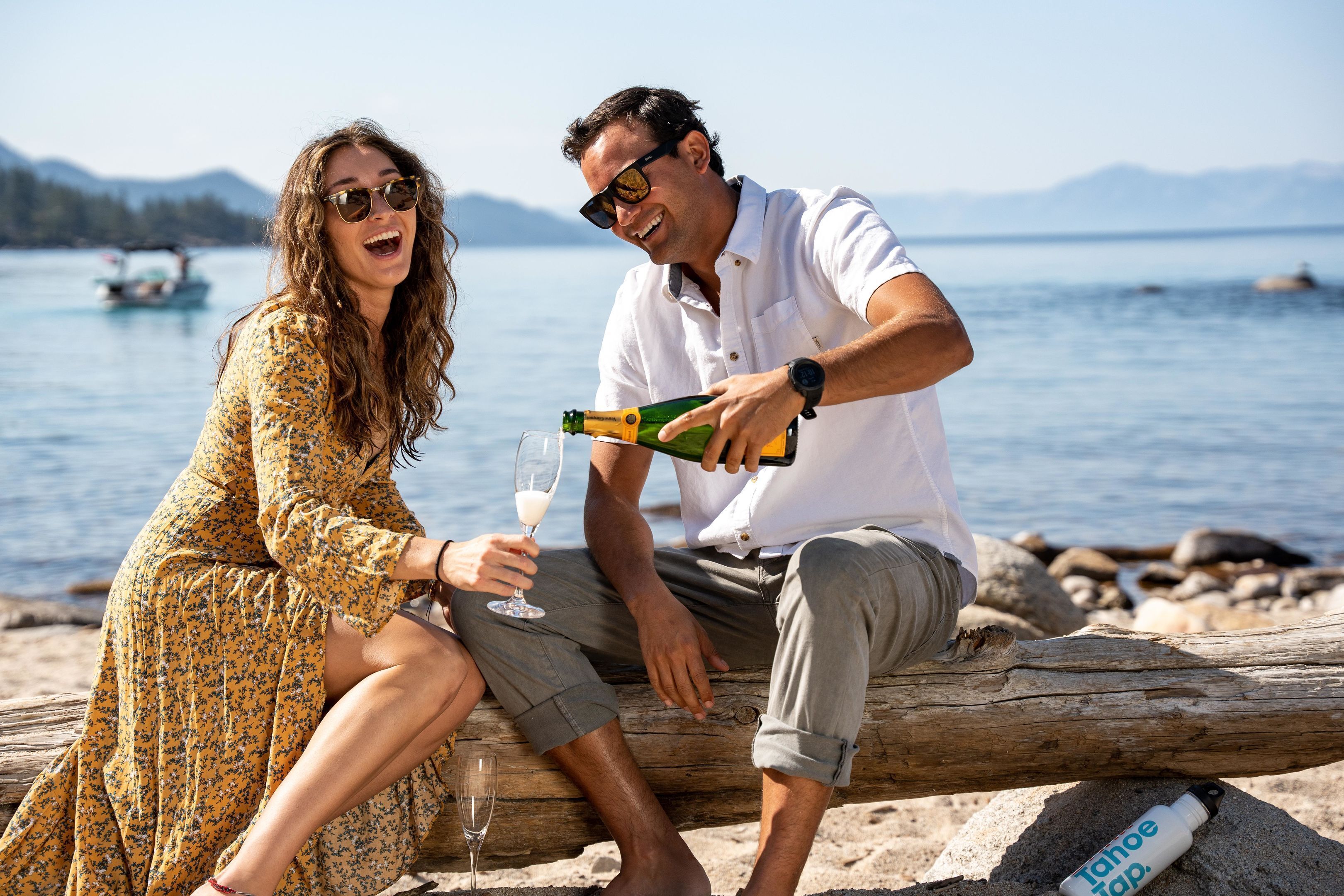 A couple sitting on a log by a lake with a bottle of champagne and a glass.