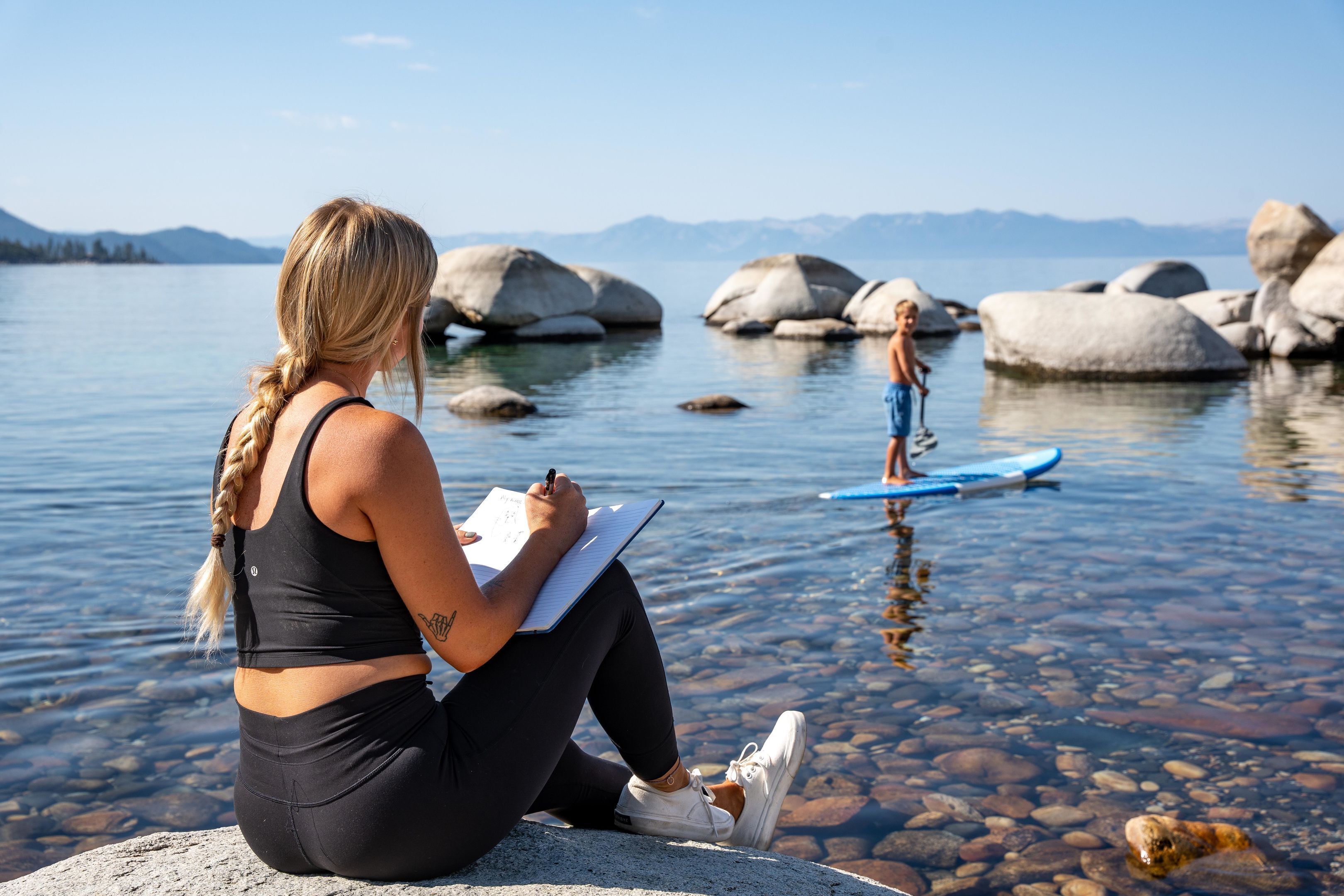 A person sitting on a rock by a lake, writing in a notebook.