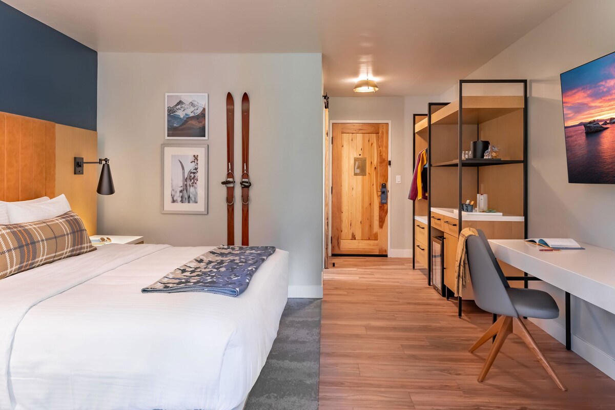 A modern hotel room with a bed, a desk, and a view of the hallway.