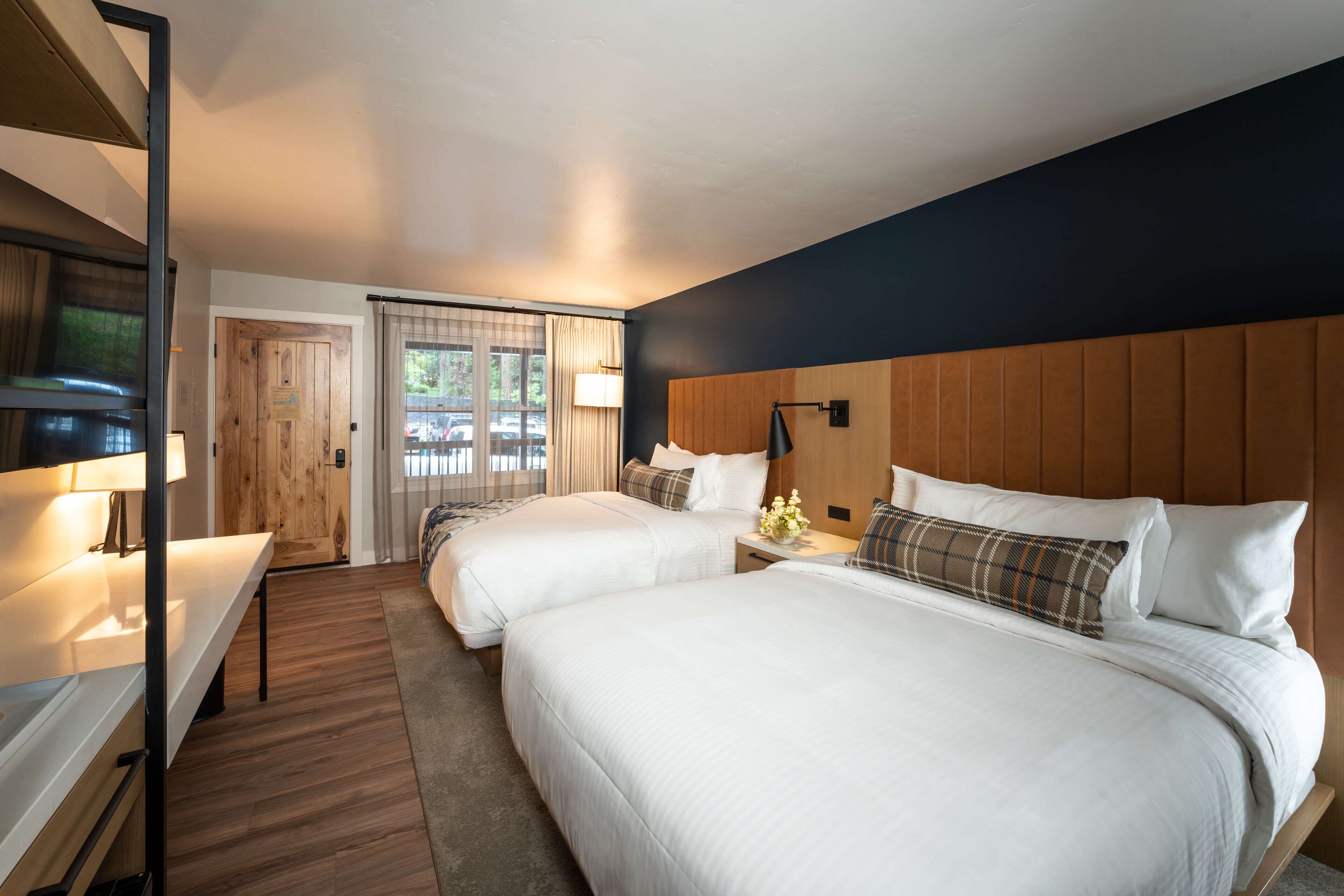 A hotel room with two beds, a wooden headboard, a desk with a lamp, and a view of the outdoors through a window.