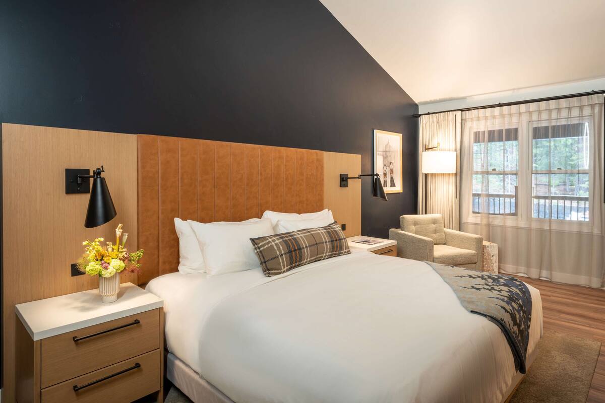 A modern hotel room with a large bed, a bedside table with a lamp, a chair, and a view of the outdoors through the window.