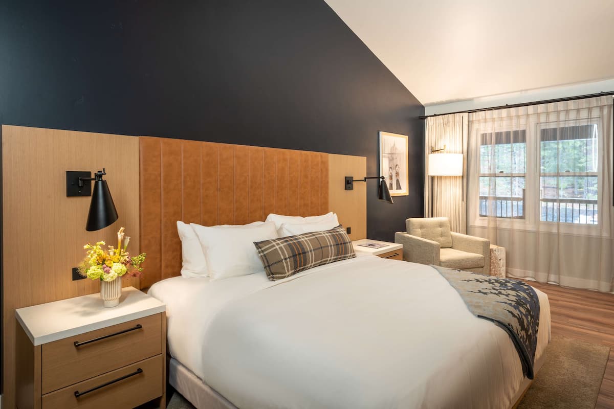 A modern hotel room with a large bed, a bedside table with a lamp, a chair, and a view of the outdoors through the window.