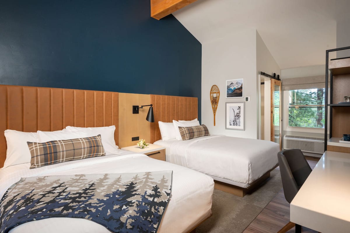 A hotel room with two beds, a wooden headboard, a small table with a lamp, and a window with a view of trees.