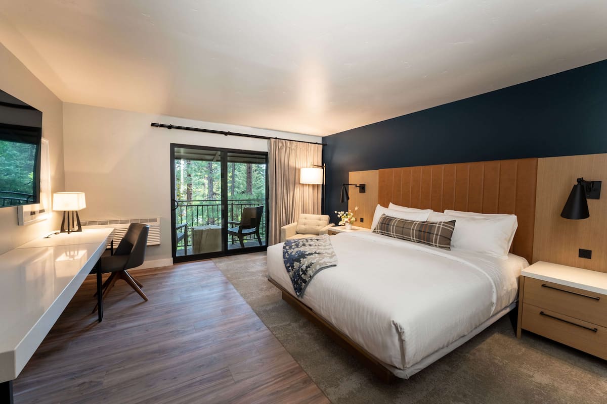 A modern hotel room with a large bed, a desk, and a chair. There is a sliding glass door leading to an outdoor area.