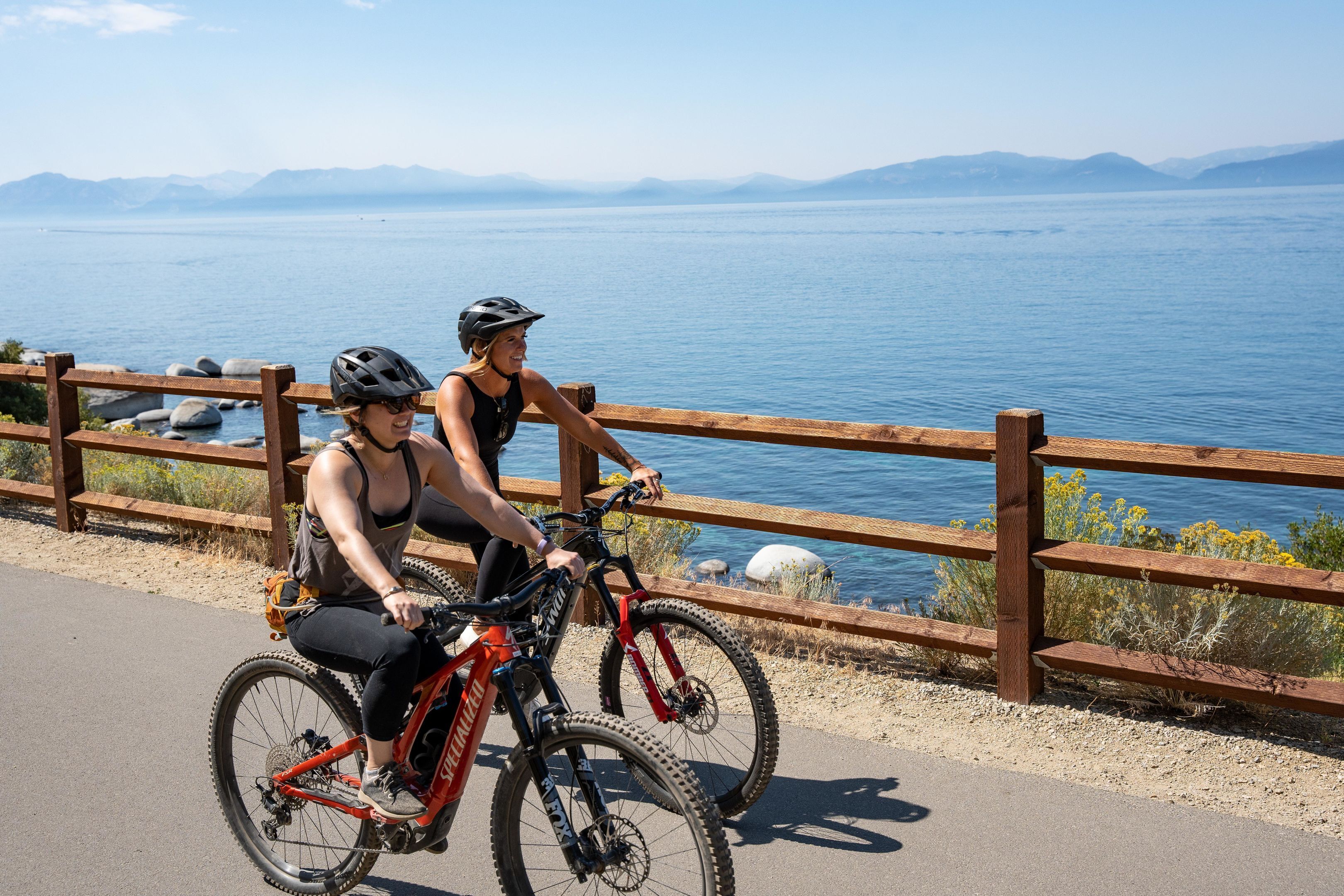 Deux personnes à vélo se trouvent près d’une clôture en bois, d’où l’on aperçoit un plan d’eau et, au loin, des montagnes.