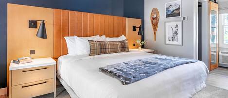 Une chambre moderne avec une tête de lit en bois, un grand lit recouvert de draps blancs, une table de chevet avec des livres et des lampes fixées au mur.