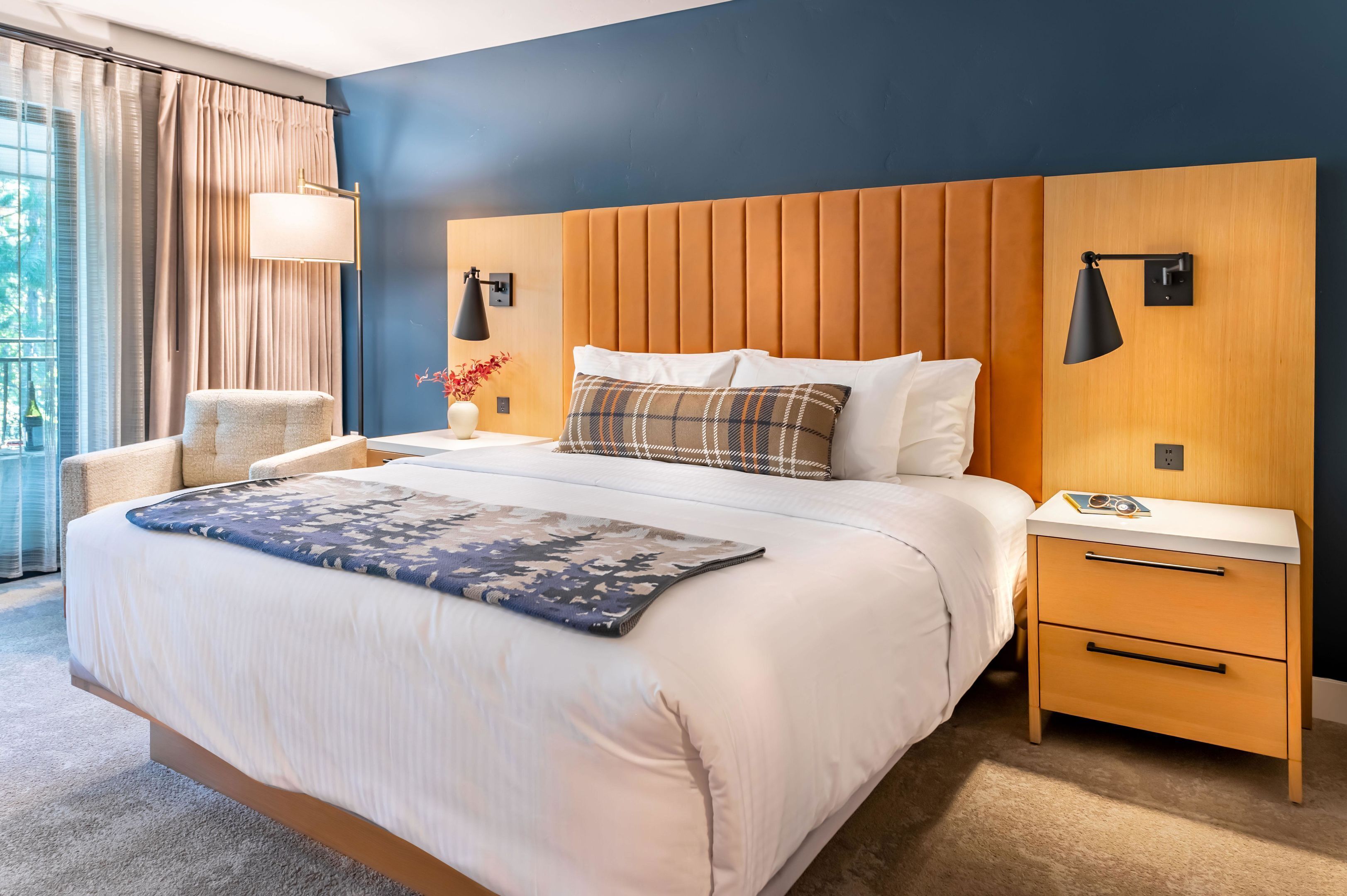 A modern hotel room with a large bed, a wooden headboard, a bedside table with a lamp, and a view of the outdoors through large windows.