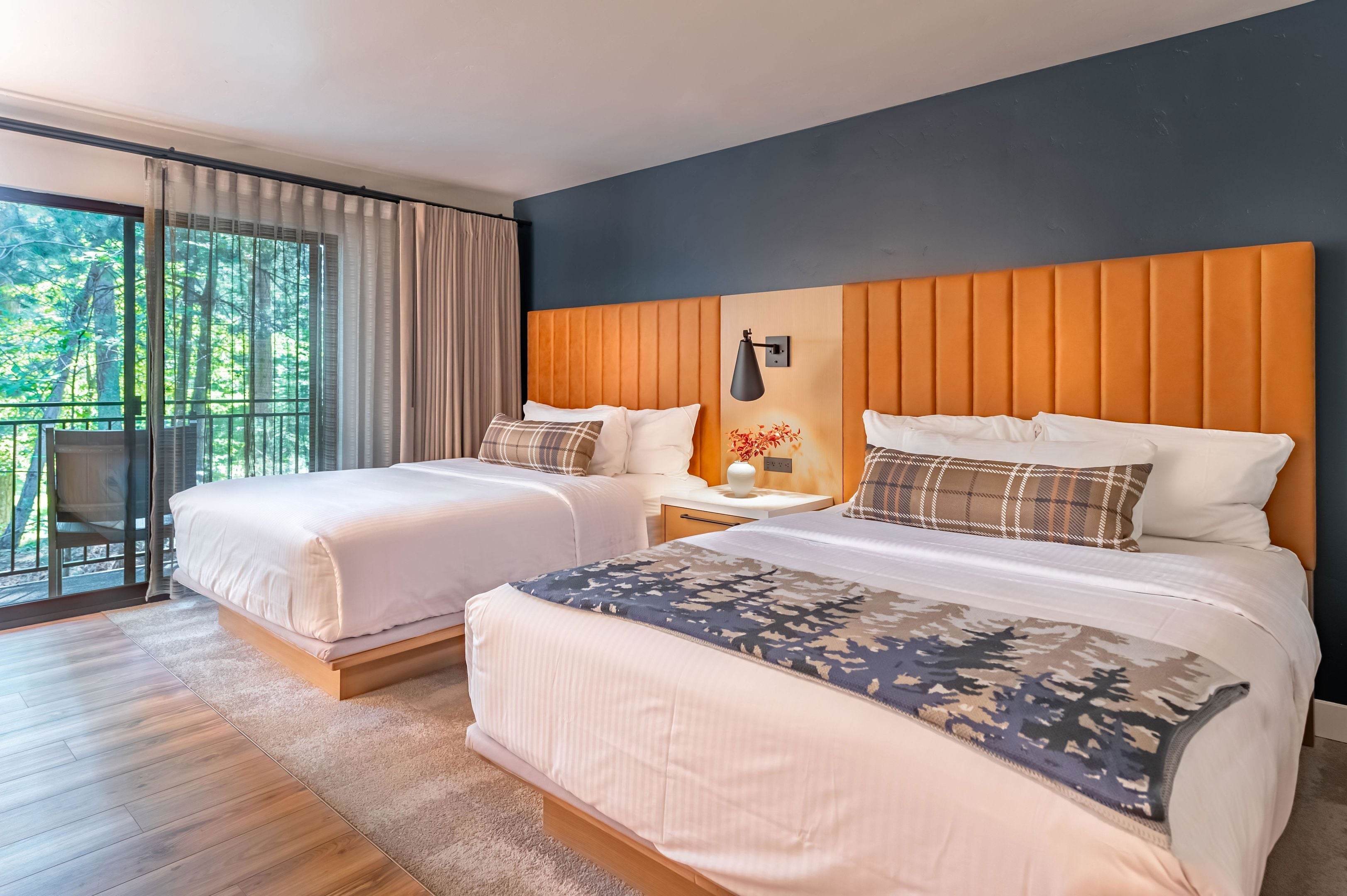 Une chambre d’hôtel avec deux lits, une grande fenêtre avec des rideaux, une table de chevet avec une lampe et une vue sur les arbres à l’extérieur.