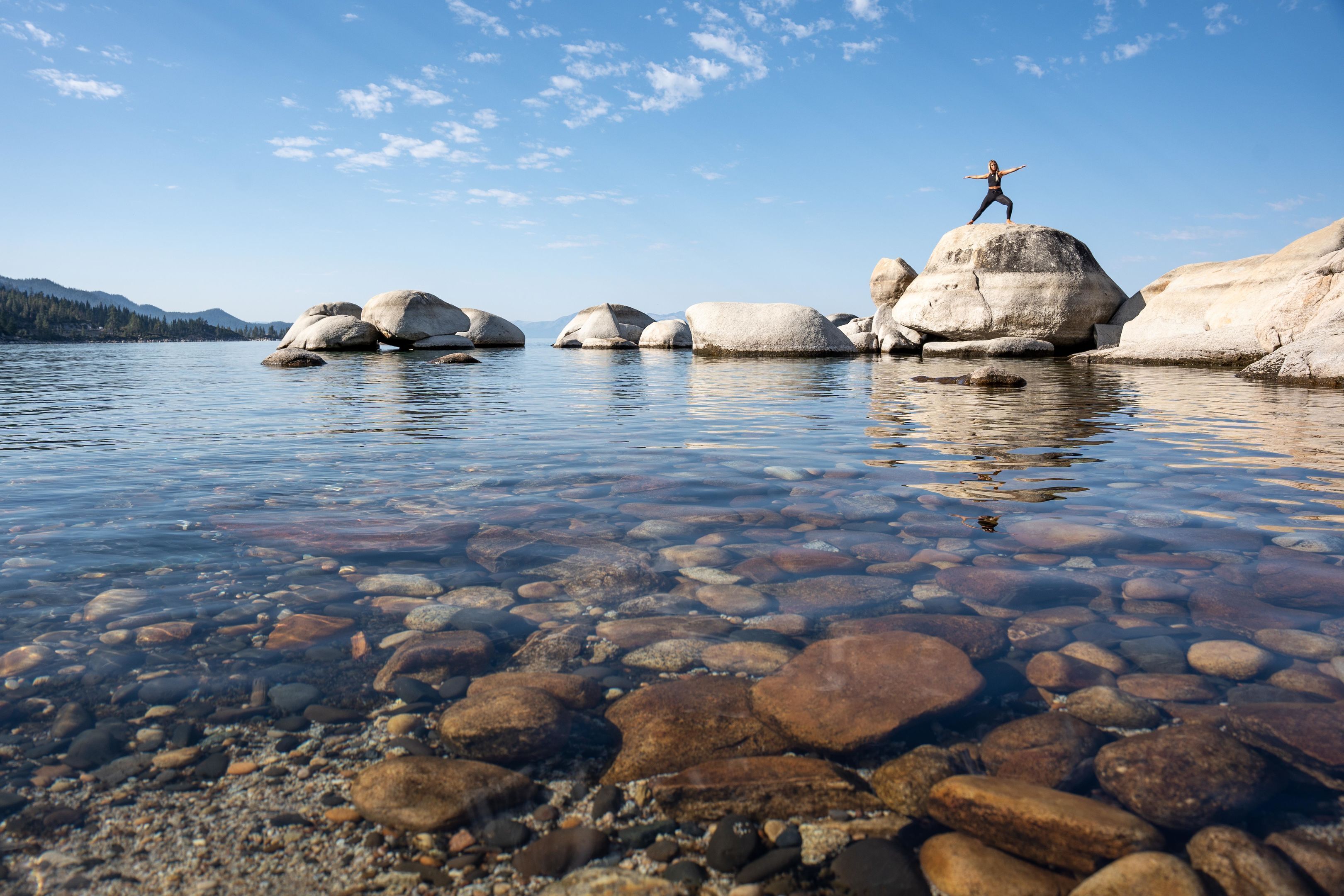 A person standing on a rock by a clear lake with visible pebbles and mountains in the background.