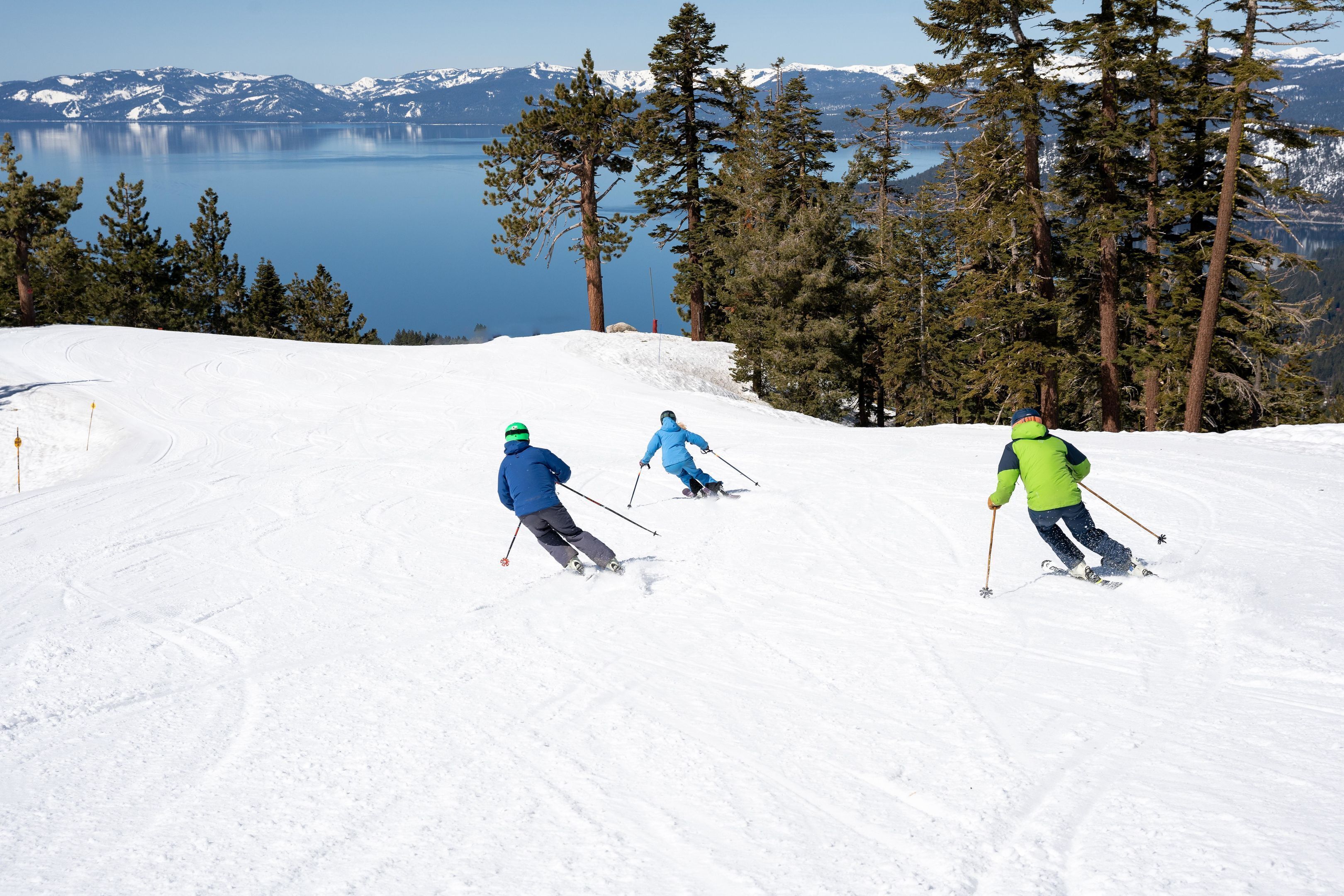 Three skiers on a snowy slope with a lake and mountains in the background.