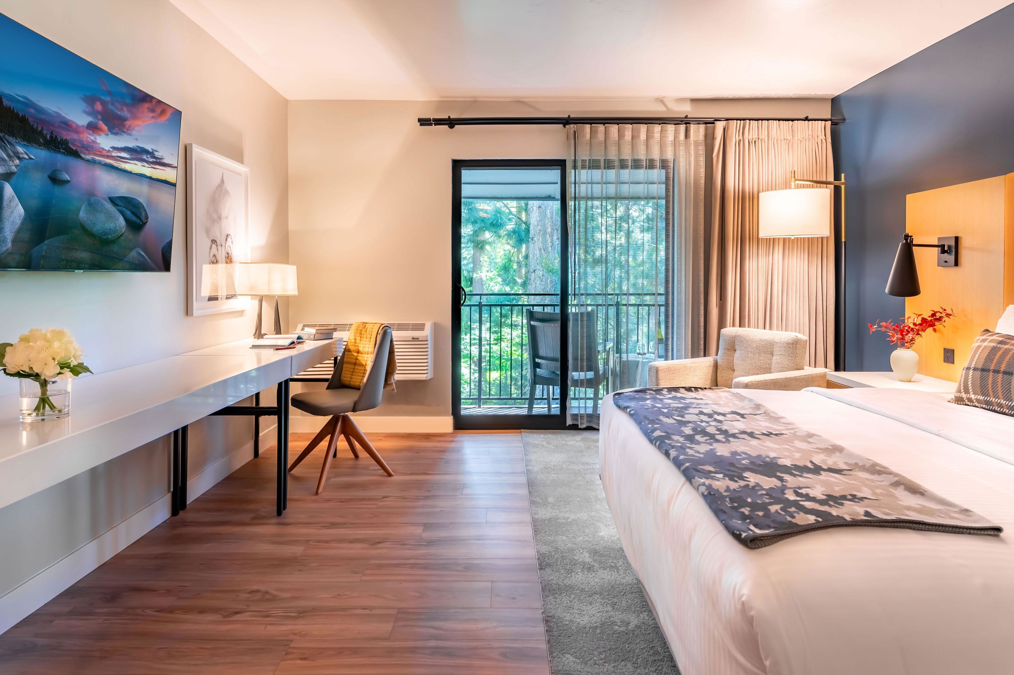 A modern hotel room with a bed, a desk, a chair, a balcony with curtains, and a painting on the wall.