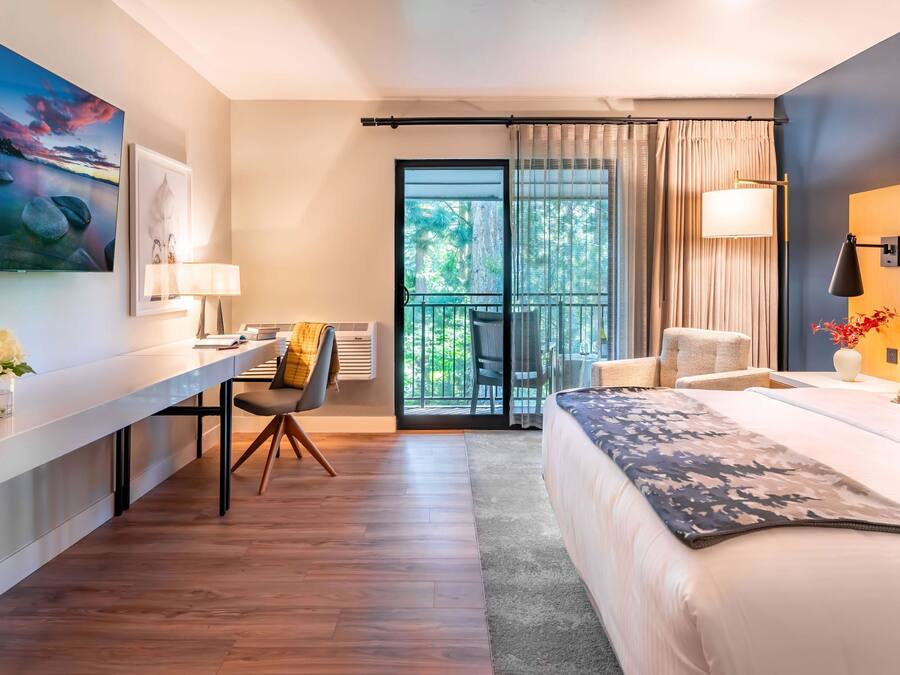A modern hotel room with a bed, a desk, a chair, a balcony with curtains, and a painting on the wall.