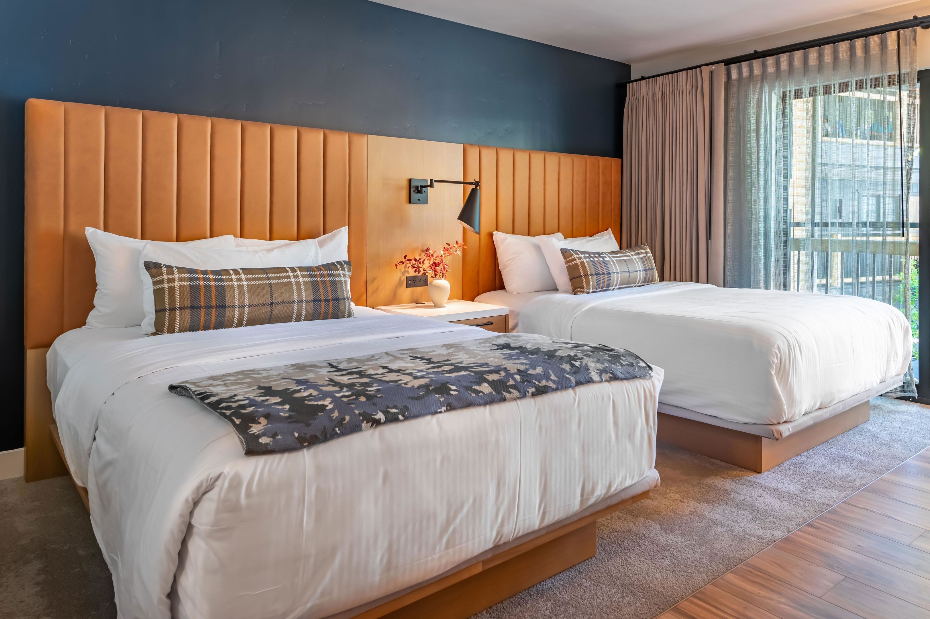 A hotel room with two beds, a wooden headboard, a nightstand with a lamp, and a view of the outdoors through large windows.