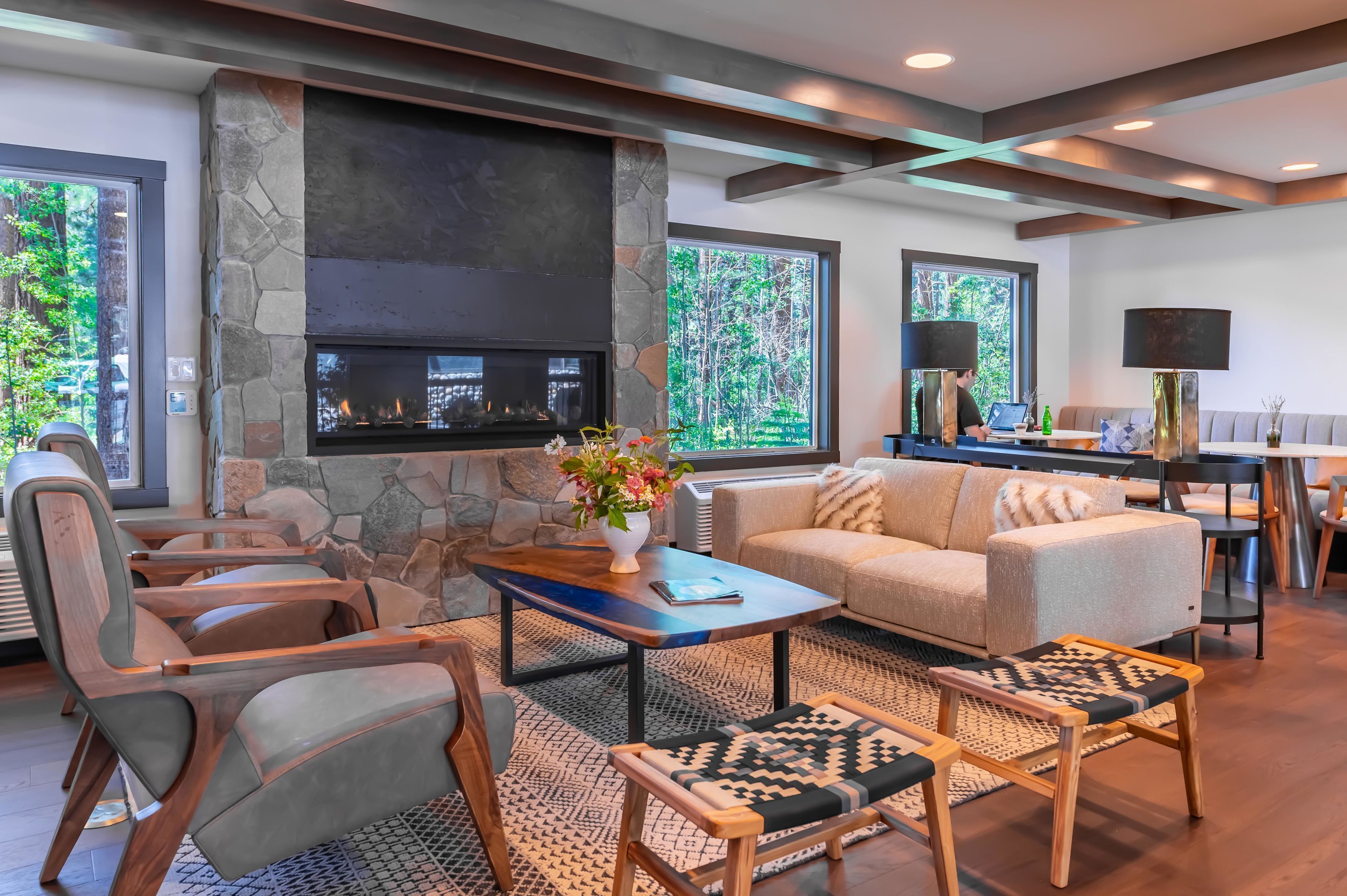 A modern living room with a stone fireplace, comfortable seating, and large windows offering a view of trees.