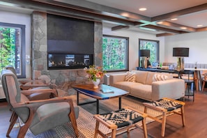 A modern living room with a stone fireplace, comfortable seating, and large windows offering a view of trees.