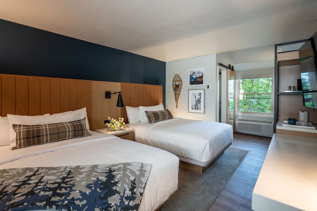 A modern hotel room with two beds, a wooden headboard, a nightstand with a lamp, and a view of greenery through a window.