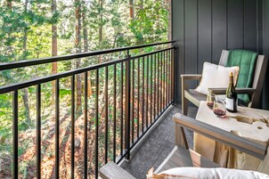 A balcony with a view of a forest, a table with a bottle and glasses, and a chair with a cushion.