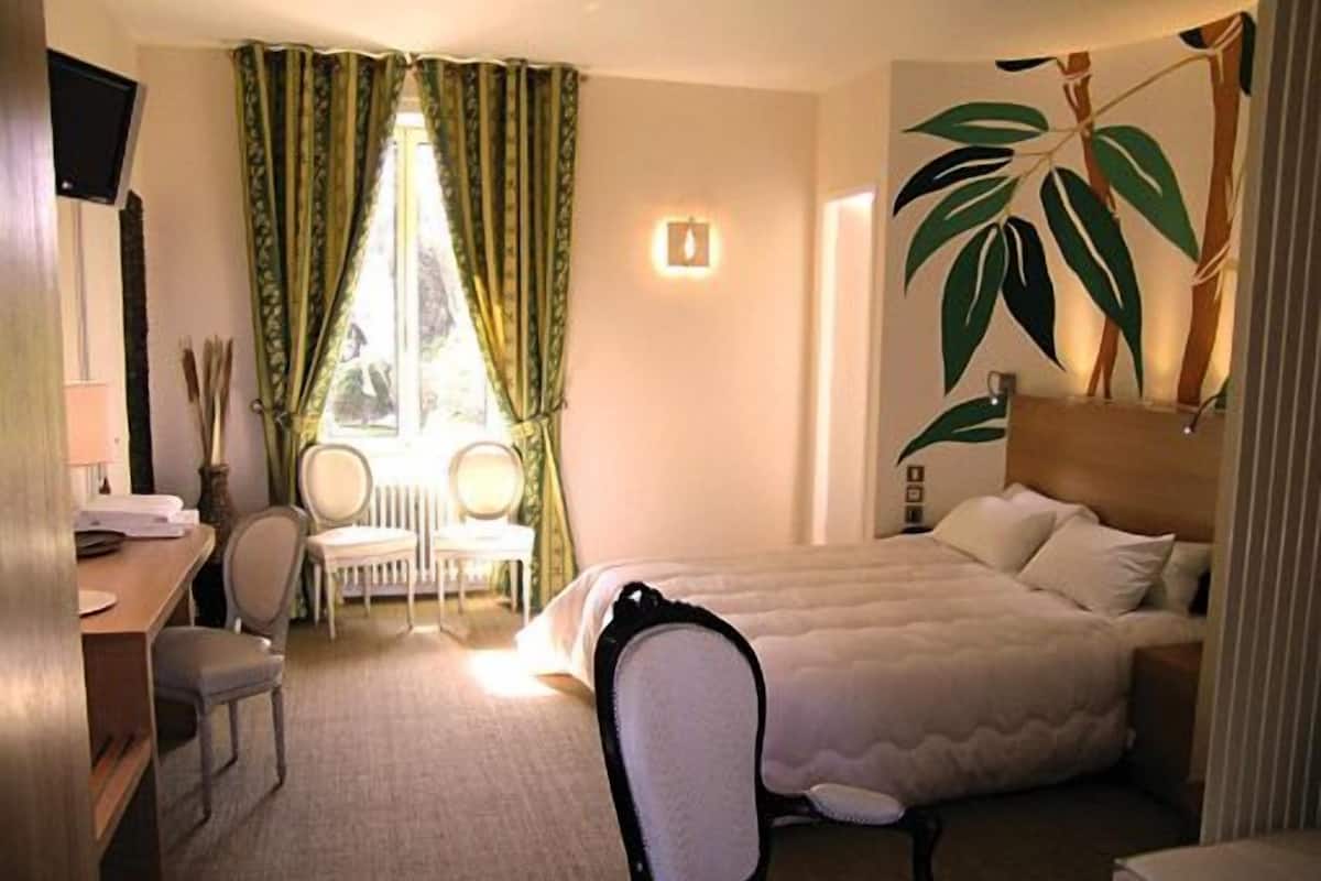 Quarto de hotel com cama, escrivaninha, cadeira e uma janela grande com cortinas.