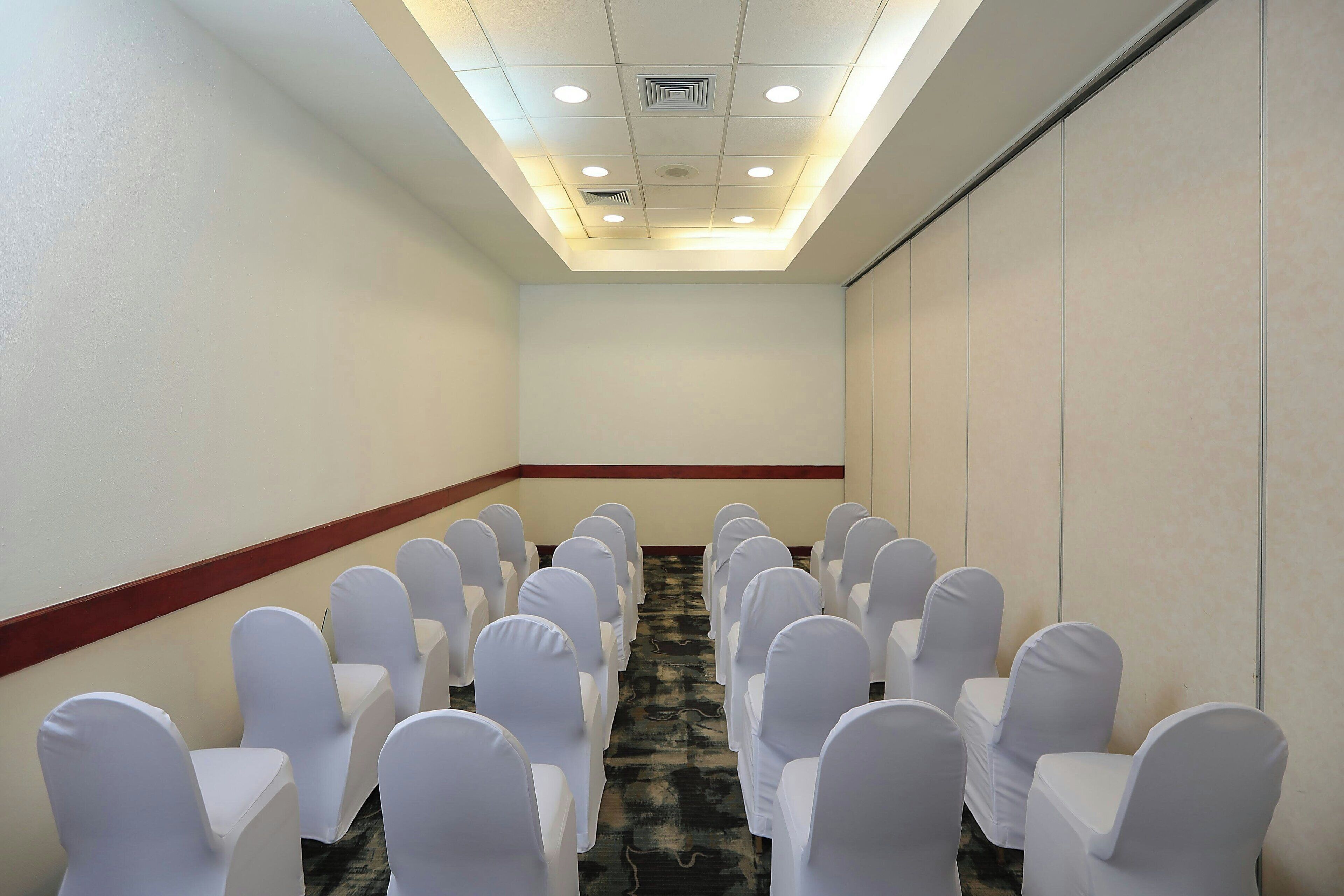 A conference room with rows of white chairs, a carpeted floor, and a ceiling with recessed lighting.