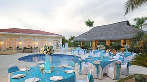 An outdoor dining area with round tables set for a formal event, surrounded by a pool and a thatched-roof structure. - Four Points by Sheraton Santo Domingo (Boca Chica)