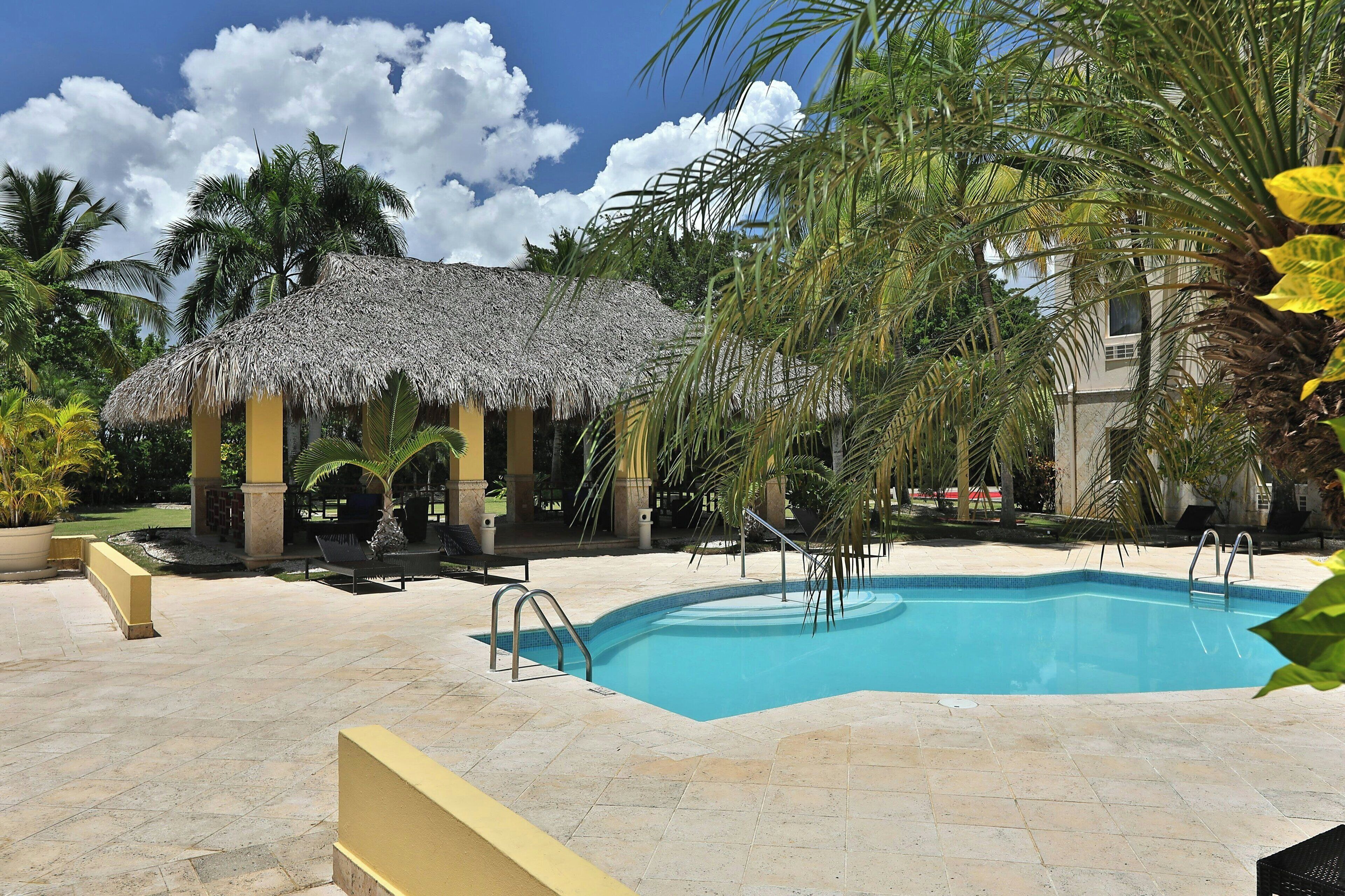 A swimming pool with a thatched-roof structure nearby, surrounded by palm trees and a building.