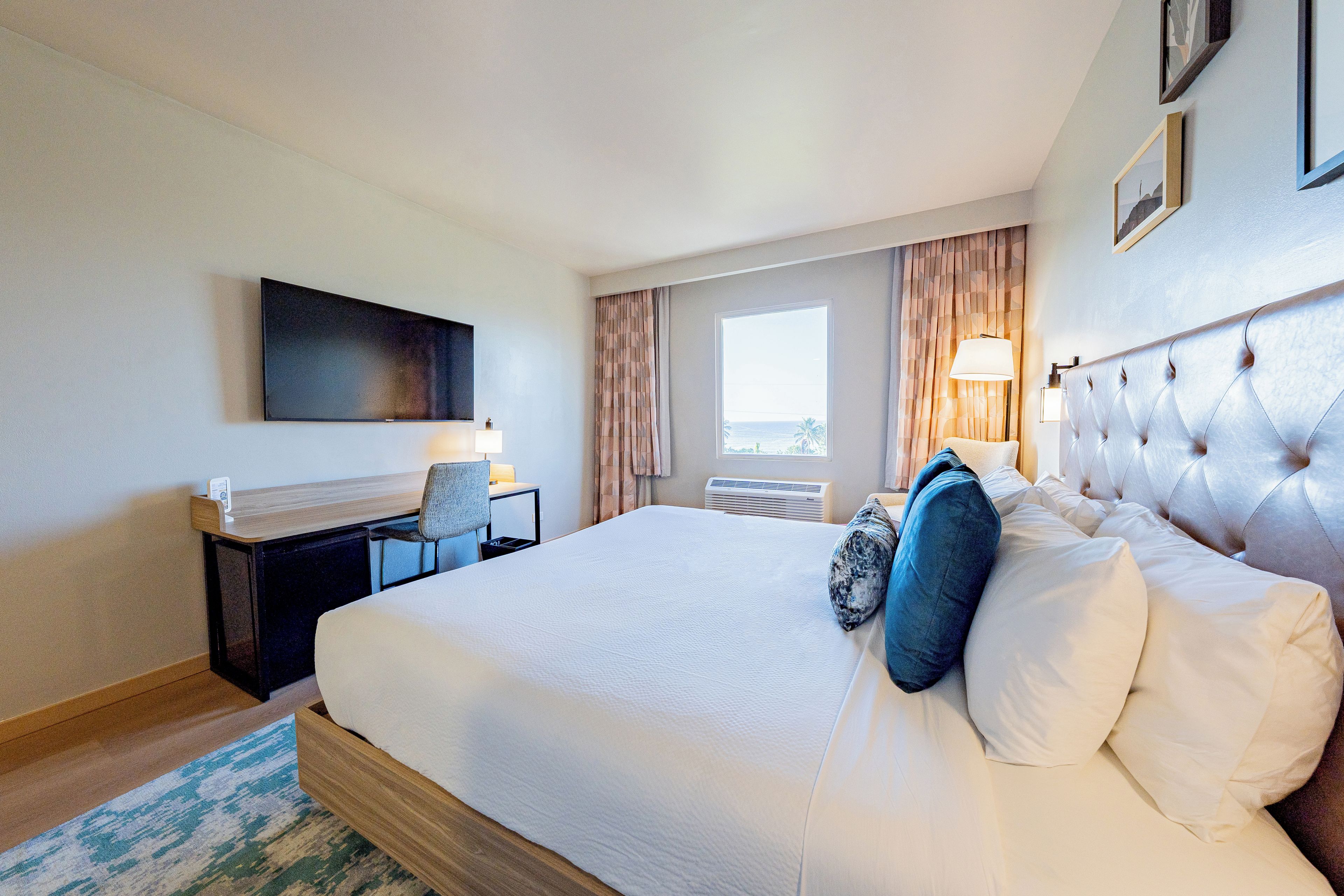 A hotel room with a large bed, a desk with a chair, a flat-screen TV mounted on the wall, a window with curtains, and a wall-mounted lamp.