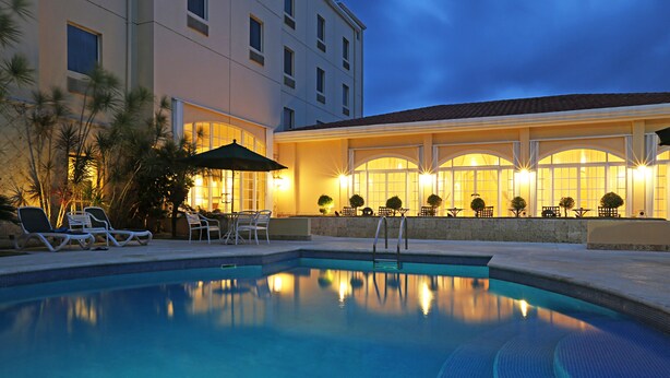 A swimming pool with lounge chairs and umbrellas, surrounded by a building with large windows and outdoor seating. - Four Points by Sheraton Santo Domingo (Boca Chica)