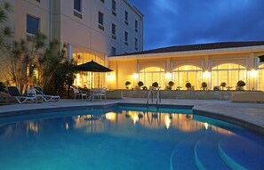 A swimming pool with lounge chairs and umbrellas, surrounded by a building with large windows and outdoor seating. - Four Points by Sheraton Santo Domingo (Boca Chica)